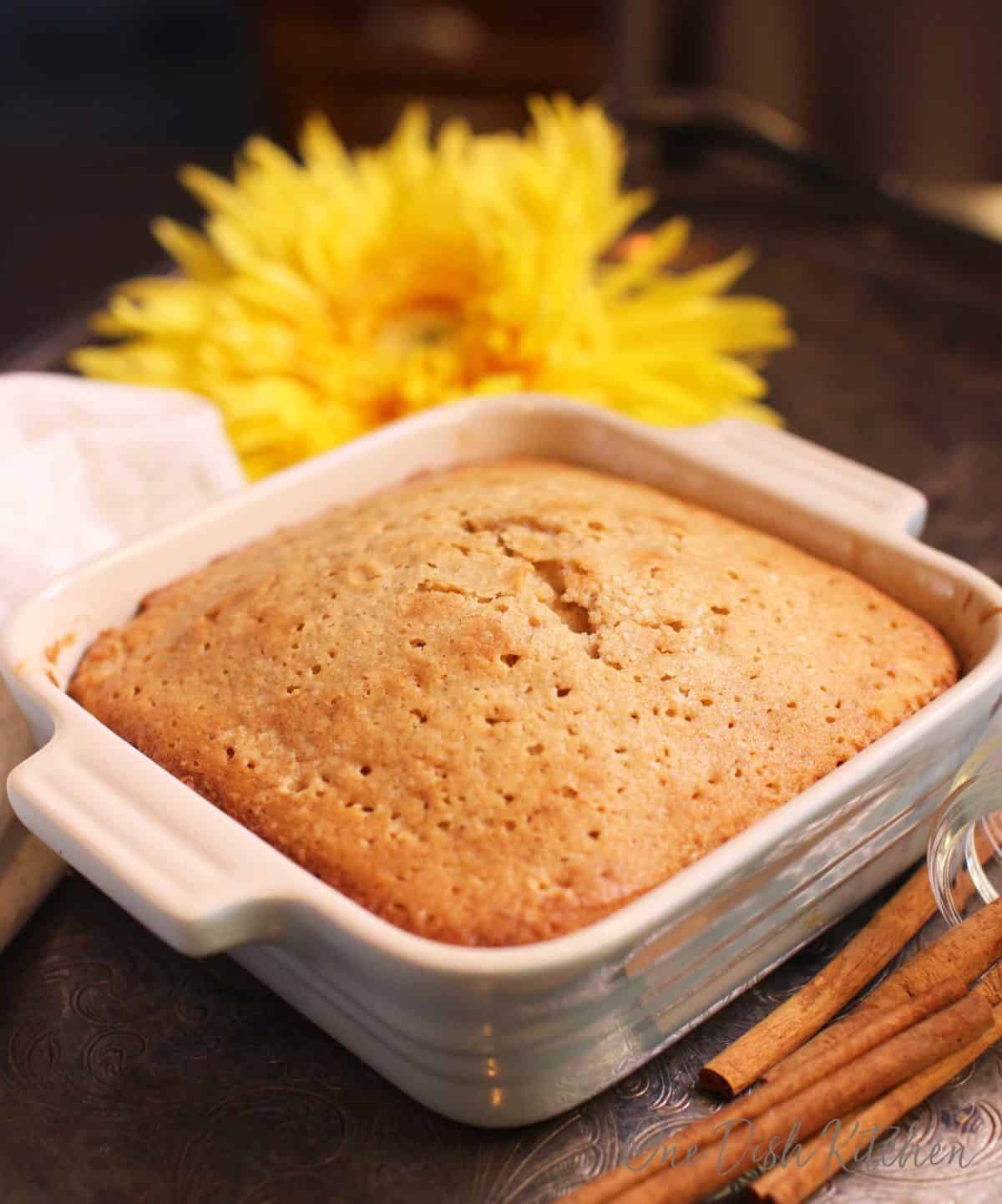 A spice cake in a small square baking dish next to yellow flowers and cinnamon sticks all on a metal tray.