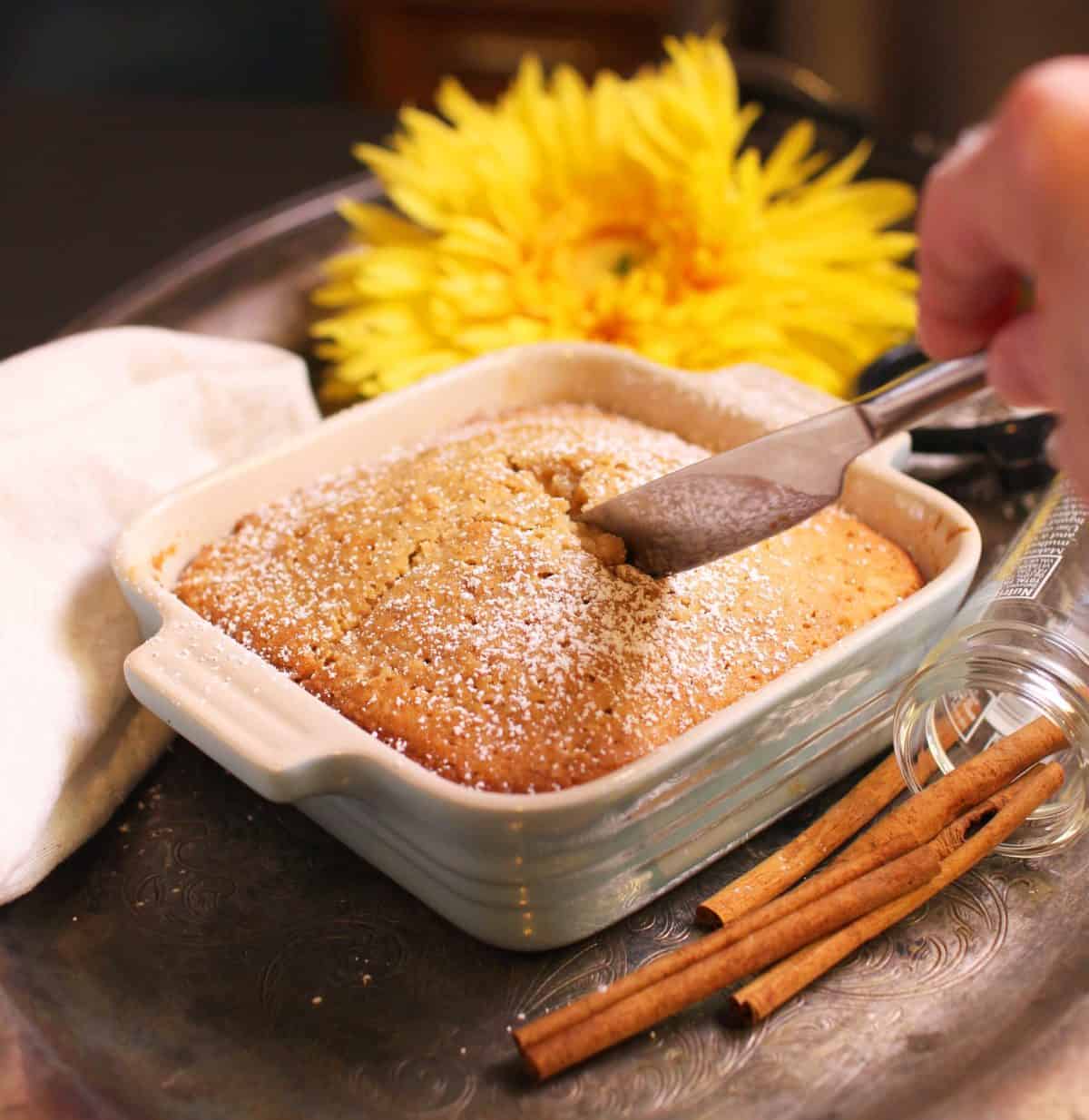 A knife cutting a piece of spice cake dusted with powdered sugar from a small baking dish on a metal tray with yellow flowers and cinnamon sticks.