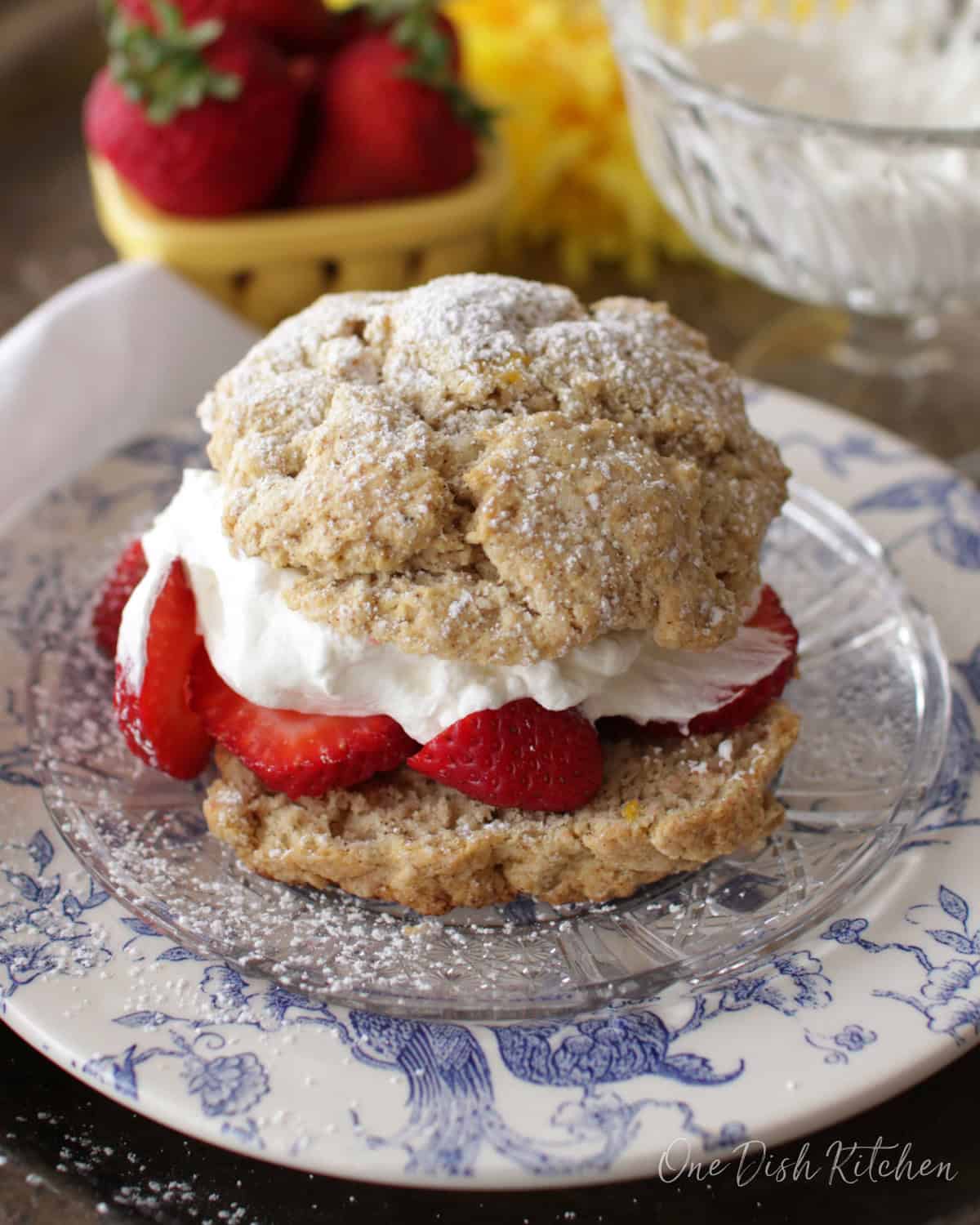 A strawberry shortcake filled with whipped cream and strawberries and dusted with powdered sugar on a plate with a bowl of whipped cream and a bowl of strawberries in the background.