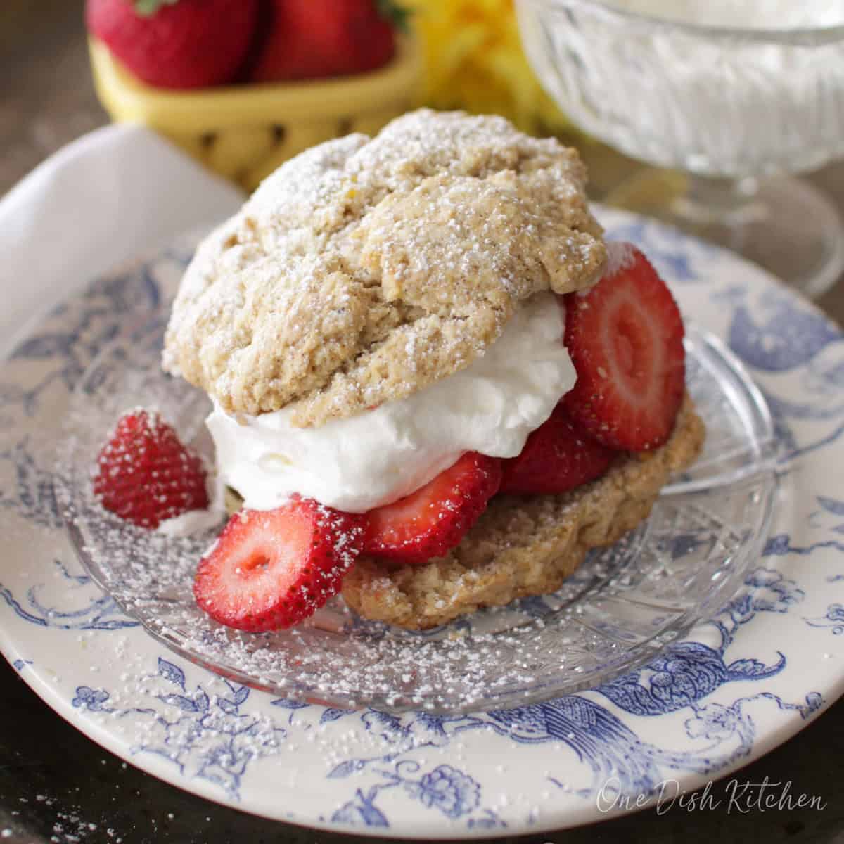 A strawberry shortcake filled with whipped cream and strawberries and dusted with powdered sugar on a plate with a bowl of whipped cream and a bowl of strawberries in the background.
