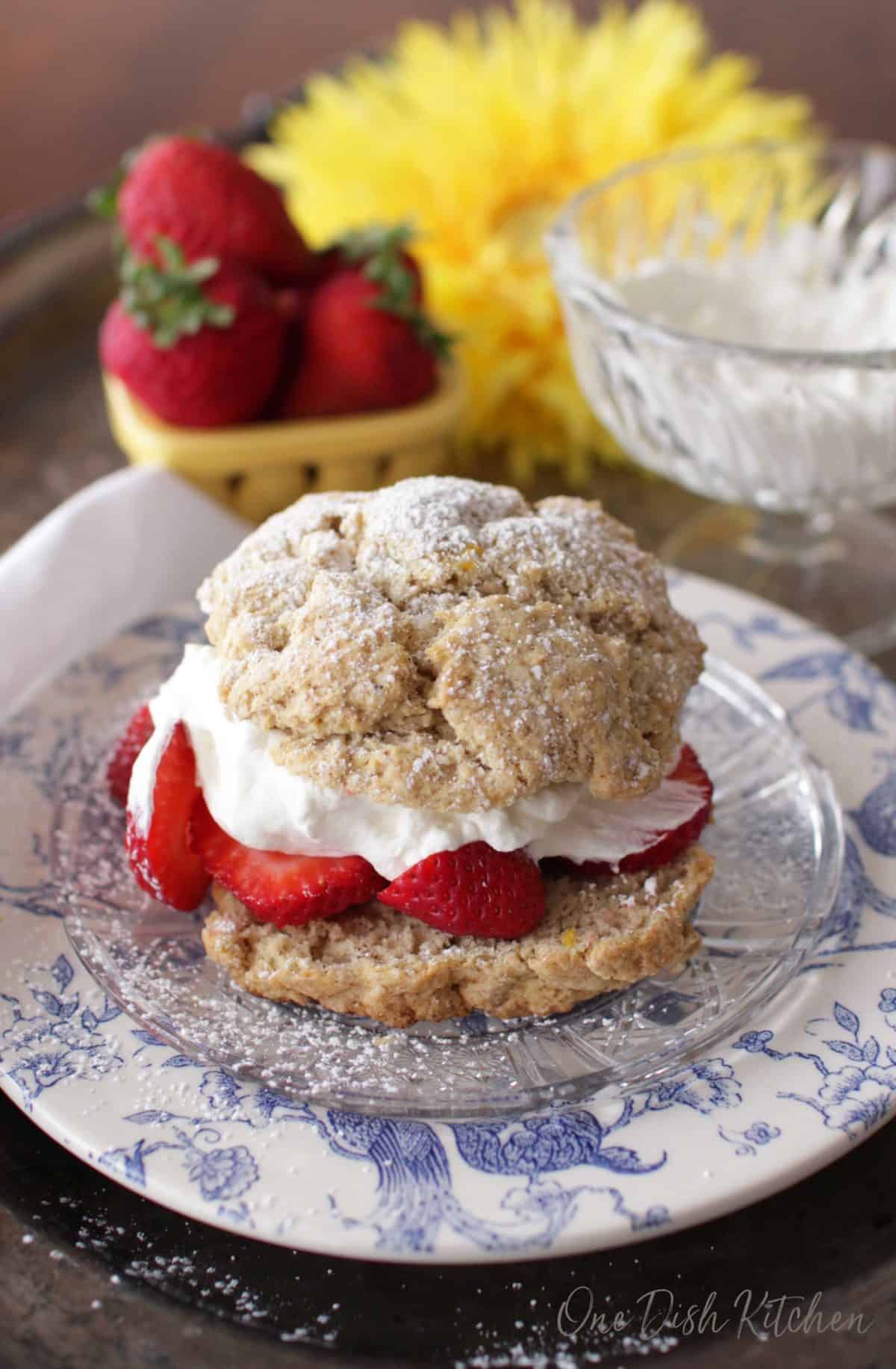 A strawberry shortcake filled with whipped cream and strawberries and dusted with powdered sugar on a plate with a bowl of whipped cream and a bowl of strawberries in the background.