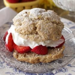 A strawberry shortcake filled with whipped cream and strawberries and dusted with powdered sugar on a plate with a bowl of whipped cream and a bowl of strawberries in the background.