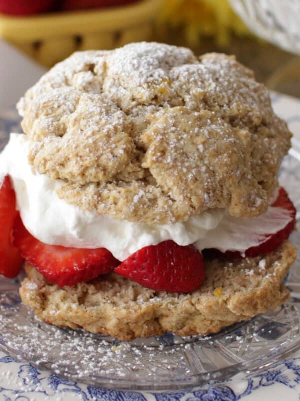 A strawberry shortcake filled with whipped cream and strawberries and dusted with powdered sugar on a plate with a bowl of whipped cream and a bowl of strawberries in the background.