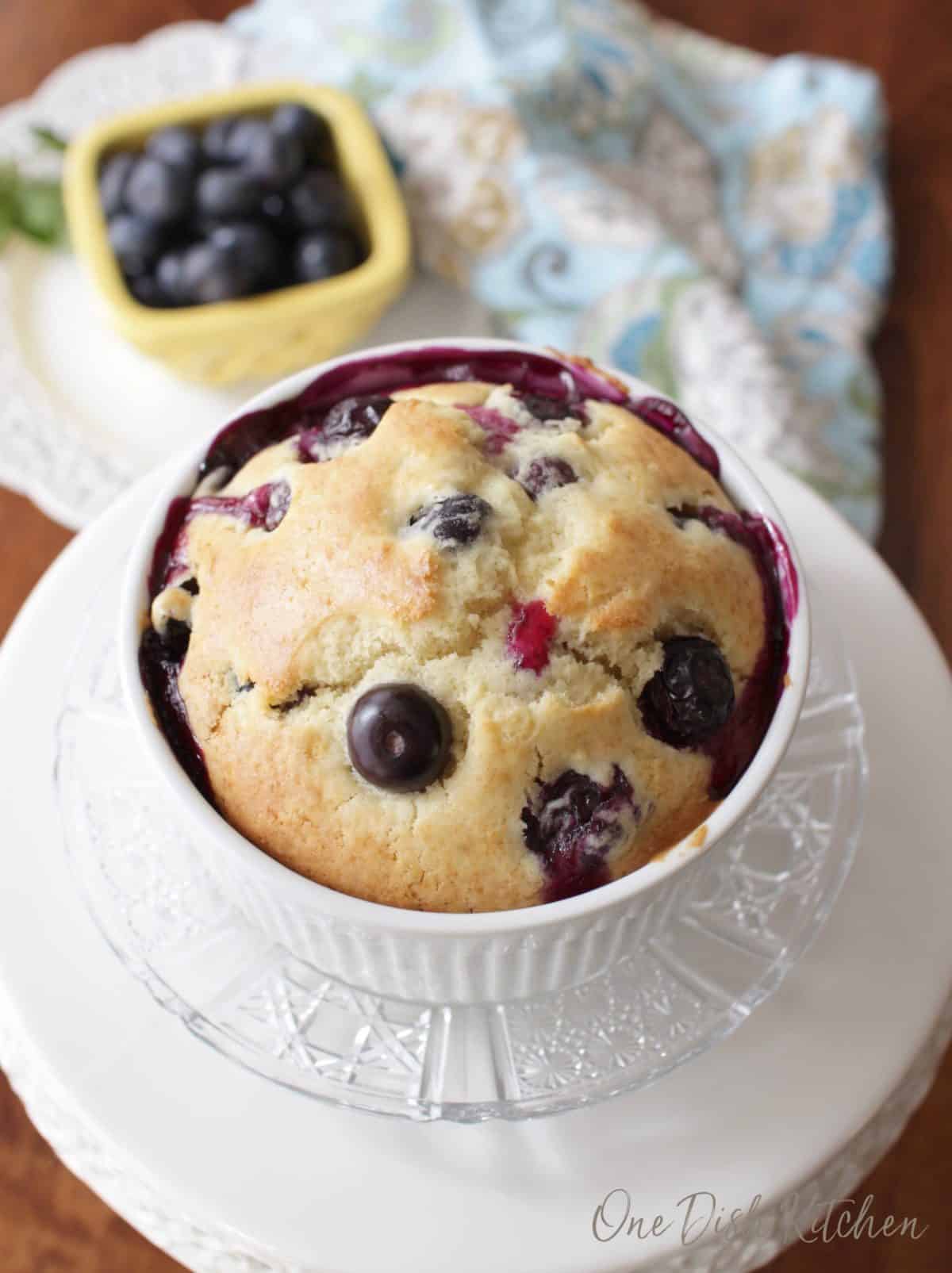 An overhead view of a blueberry muffin in a ramekin plated next to a small bowl of blueberries.