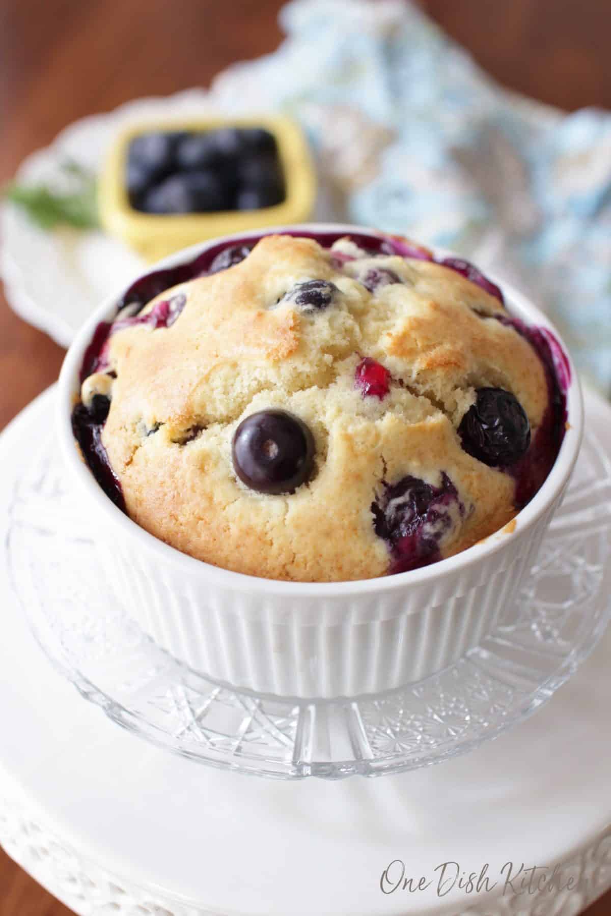 A blueberry muffin in a ramekin plated next to a small bowl of blueberries.
