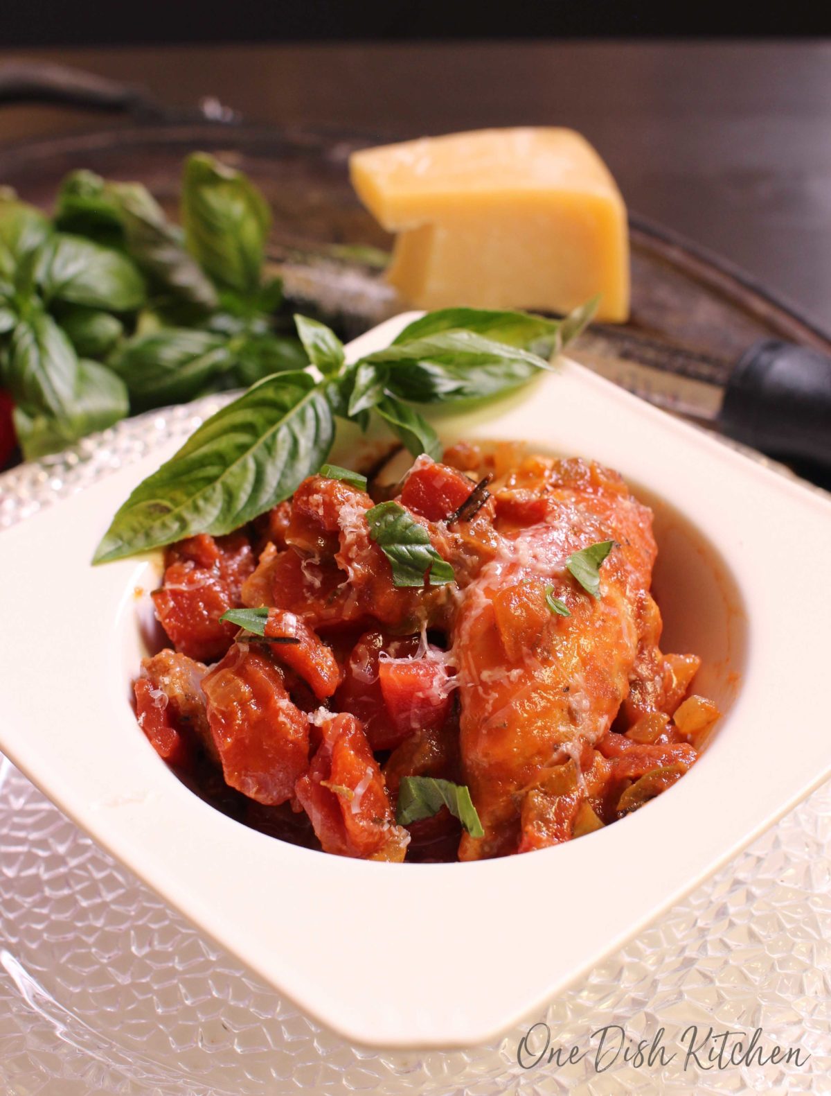 A bowl of chicken cacciatore topped with fresh basil on a metal tray with a wedge of parmesan and a cheese grater in the background.
