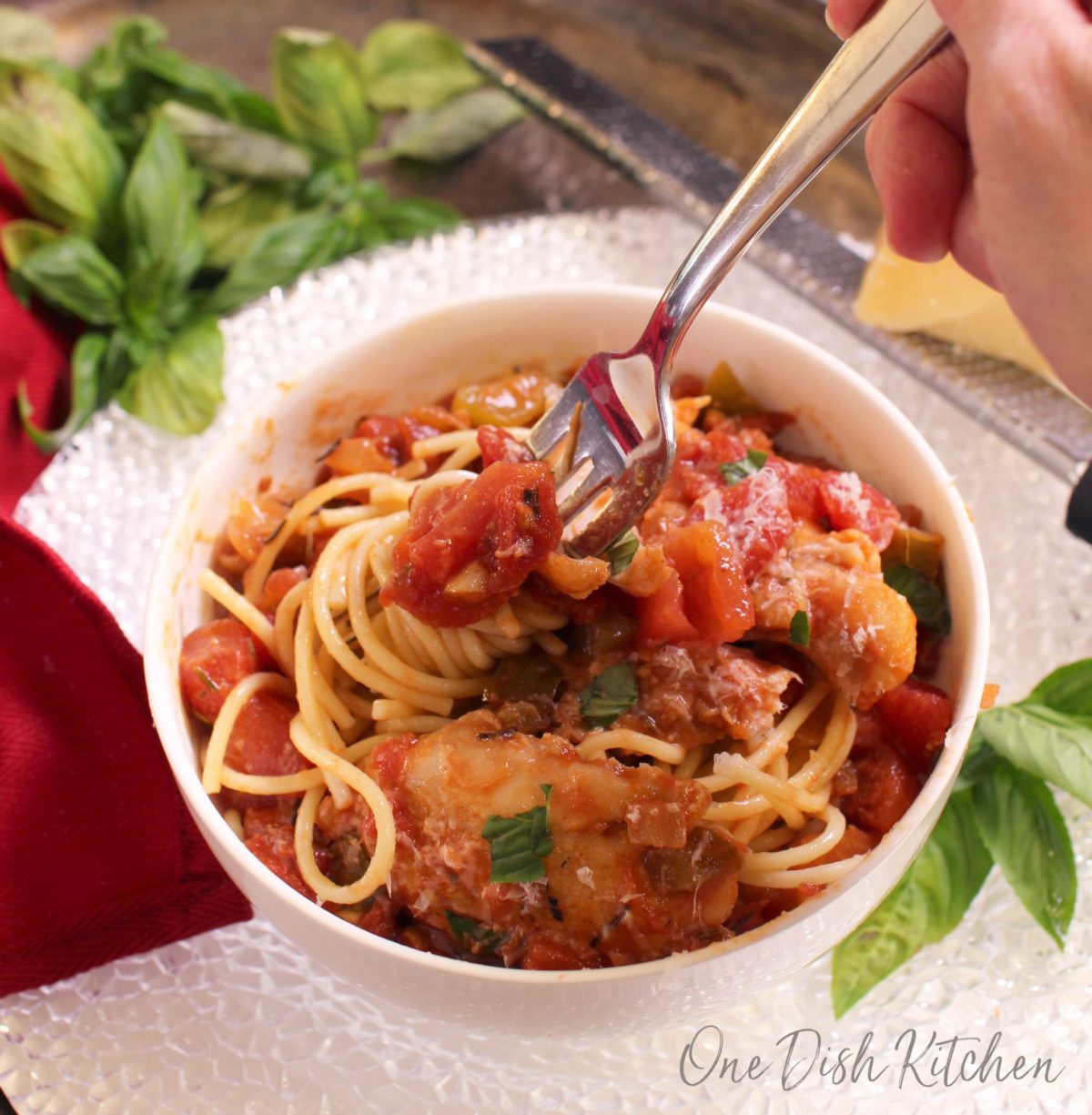 An overhead view of a bowl of chicken cacciatore with pasta and a fork swirling the pasta around the tines with scattered basil leaves in the background