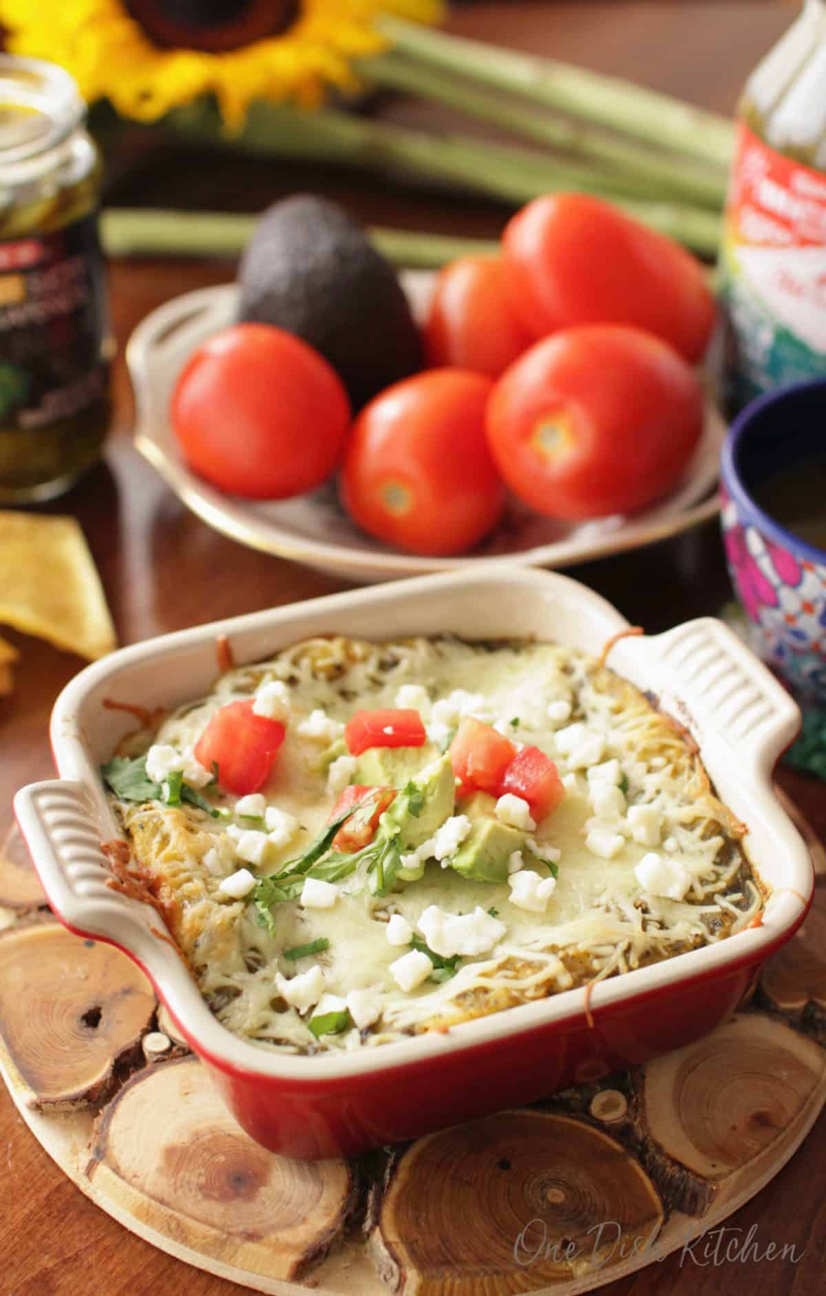 Enchiladas topped with crumbled cotija cheese and chopped avocado and tomatoes in a small baking dish on a wooden trivet next to a bowl of tomatoes and avocados.