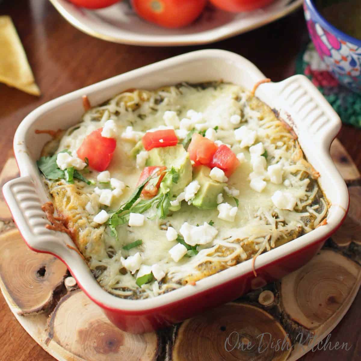 Enchiladas topped with crumbled cotija cheese and chopped avocado and tomatoes in a small baking dish on a wooden trivet.