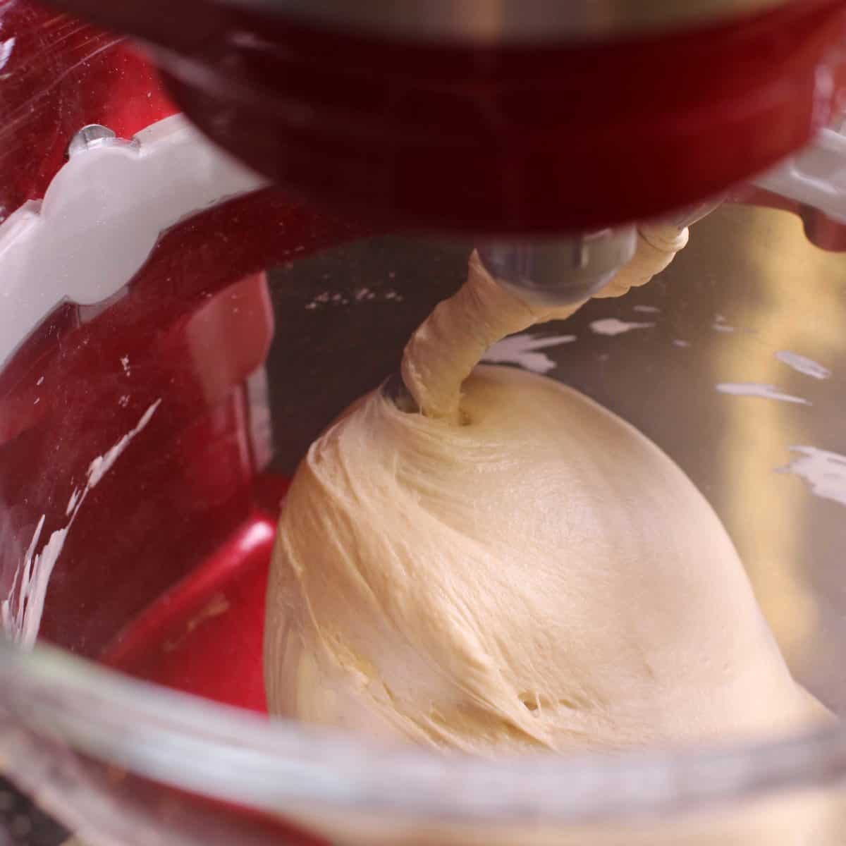 bread dough in a red electric stand mixer being mixed with a dough hook.