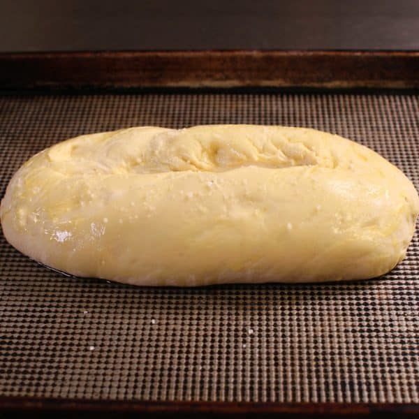 An unbaked loaf of bread on a baking sheet topped with olive oil and kosher salt.
