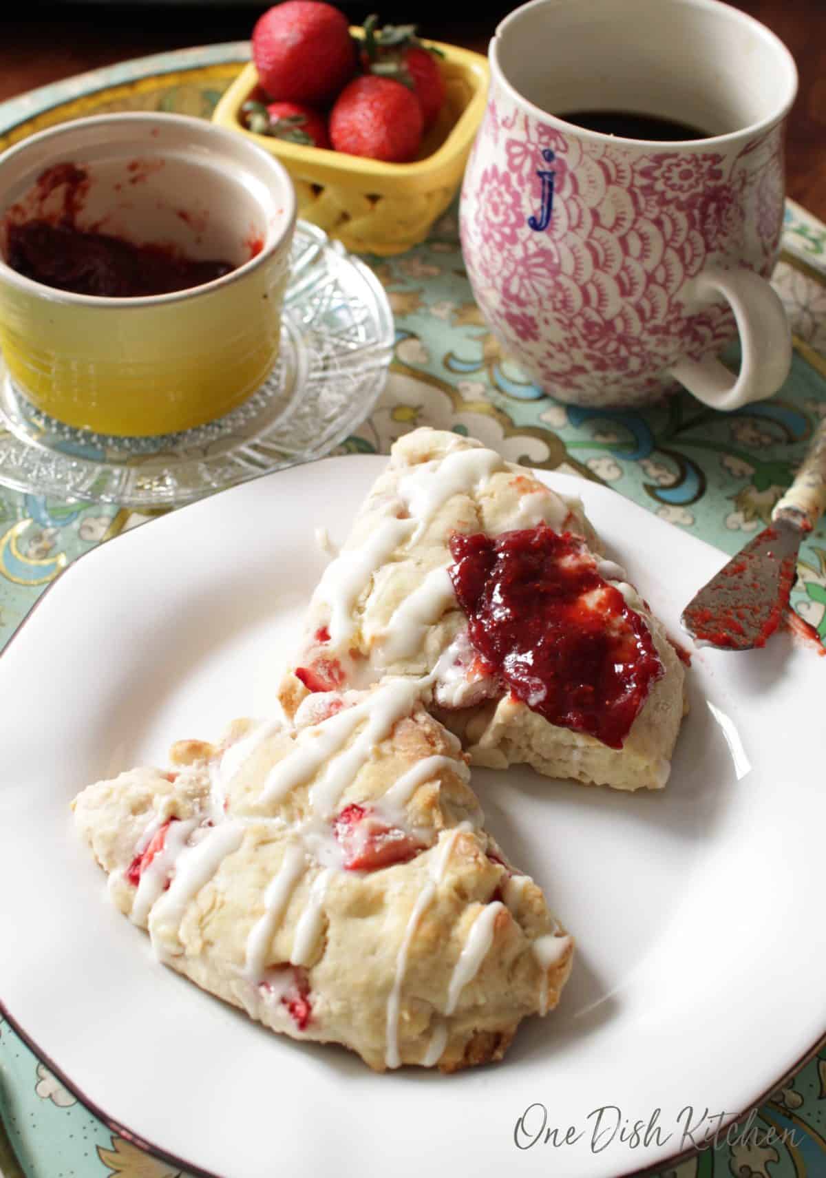 Strawberry jam spread on a scone that is on a plate next to a jar of jam, a cup of coffee, and a small bowl of strawberries all on a metal tray.