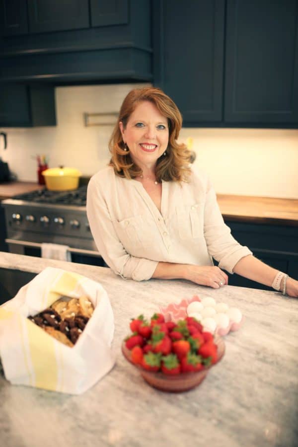 Joanie Zisk standing in the kitchen next to a counter and a basket of cookies and strawberries.