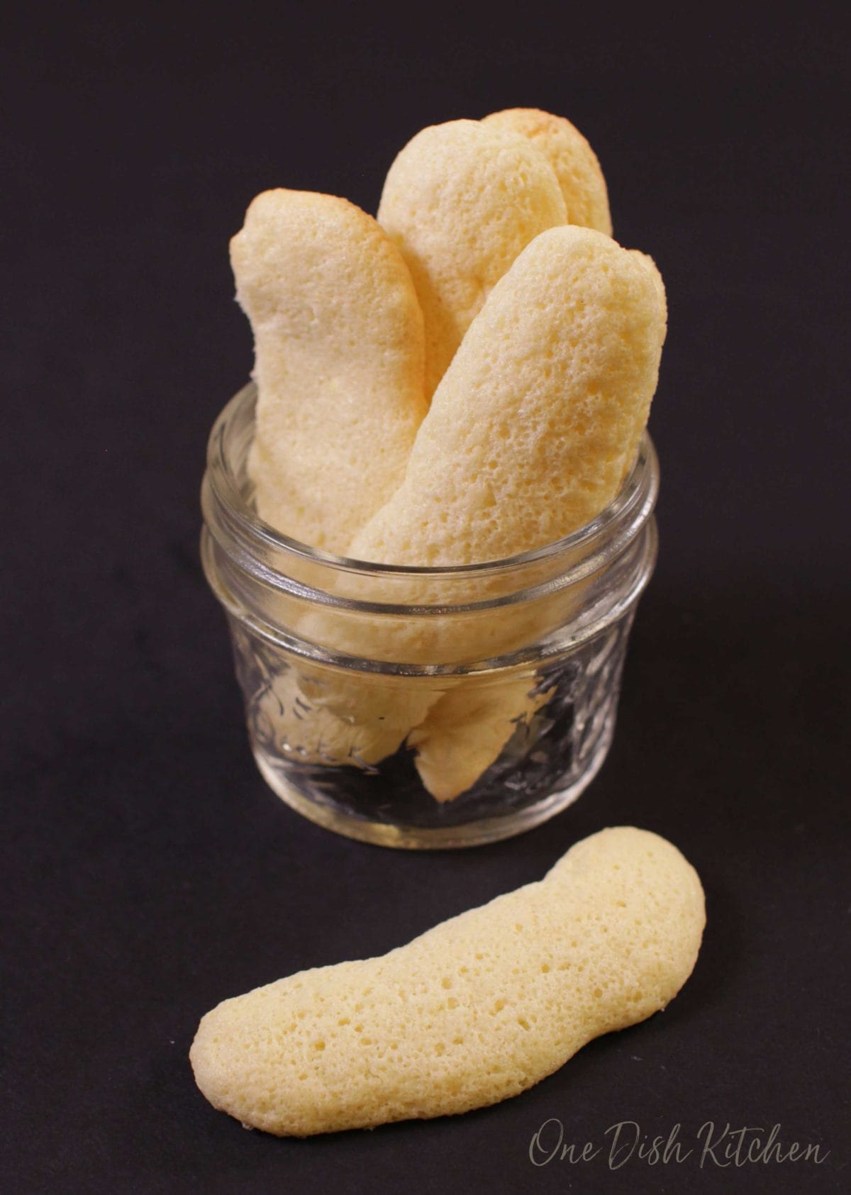 A small jar of homemade ladyfingers with one laying on a black table.