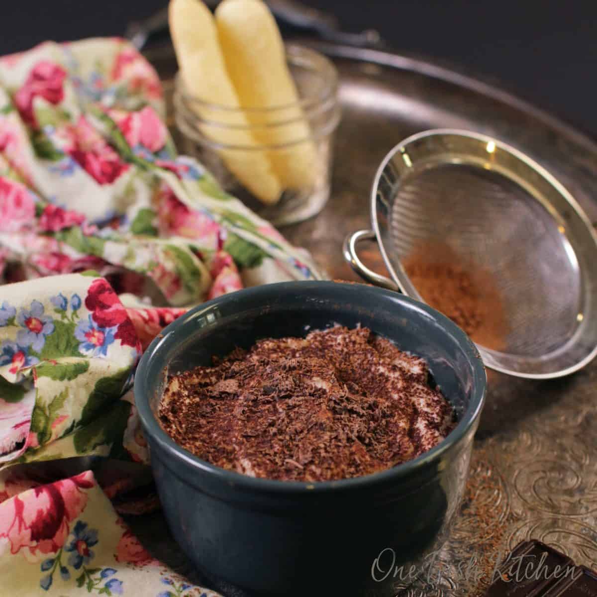 Tiramisu in a small circular dish topped with chocolate shavings next to a jar of homemade ladyfingers and a floral cloth napkin all on a metal tray