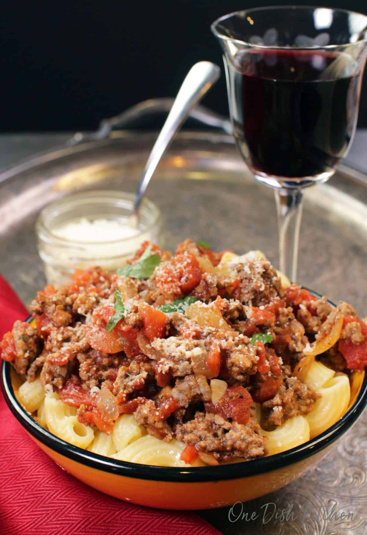 A bowl of meat sauce with pasta on a tray next to a tall glass of red wine.