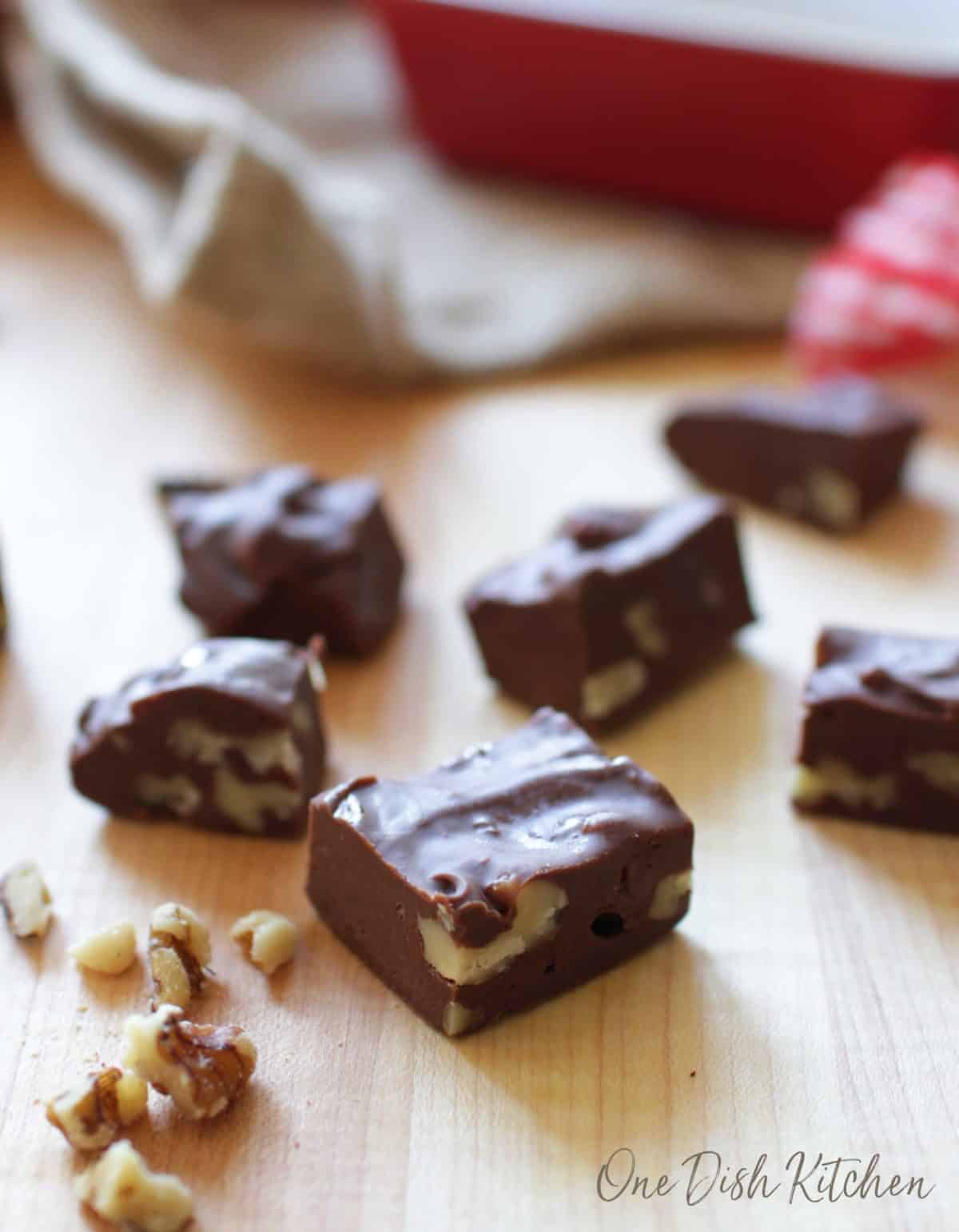 Seven squares of fudge on a brown table with scattered walnuts around the fudge.