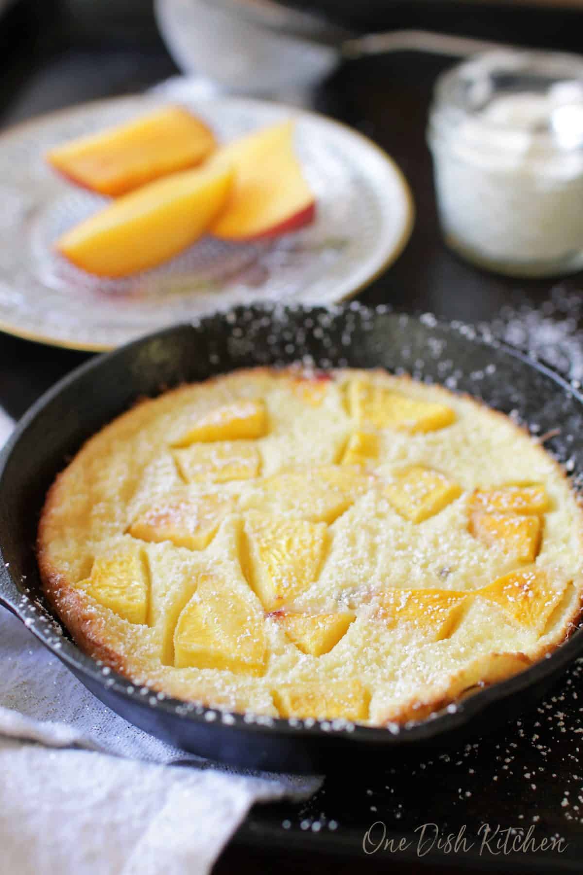 A puffed pancake in a small cast iron skillet topped with chopped peaches and dusted with powdered sugar with a small jar of whipped cream and a plate of sliced peaches in the background