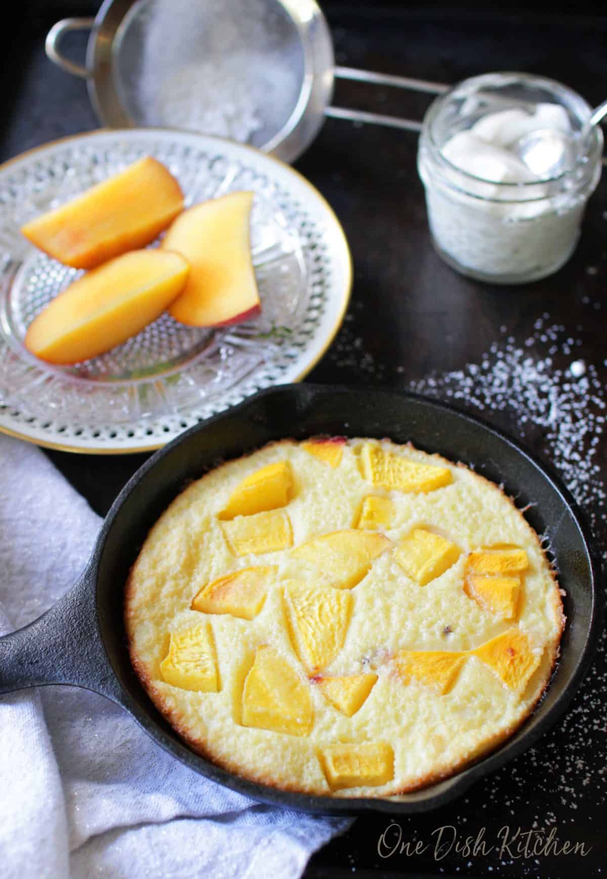 An overhead view of a peach filled dutch baby next to a small jar of whipped cream and a plate of sliced peaches 