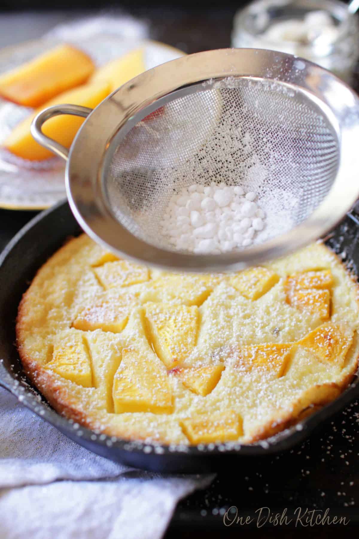 A peach filled puffed pancake in a small cast iron skillet being dusted with powdered sugar.