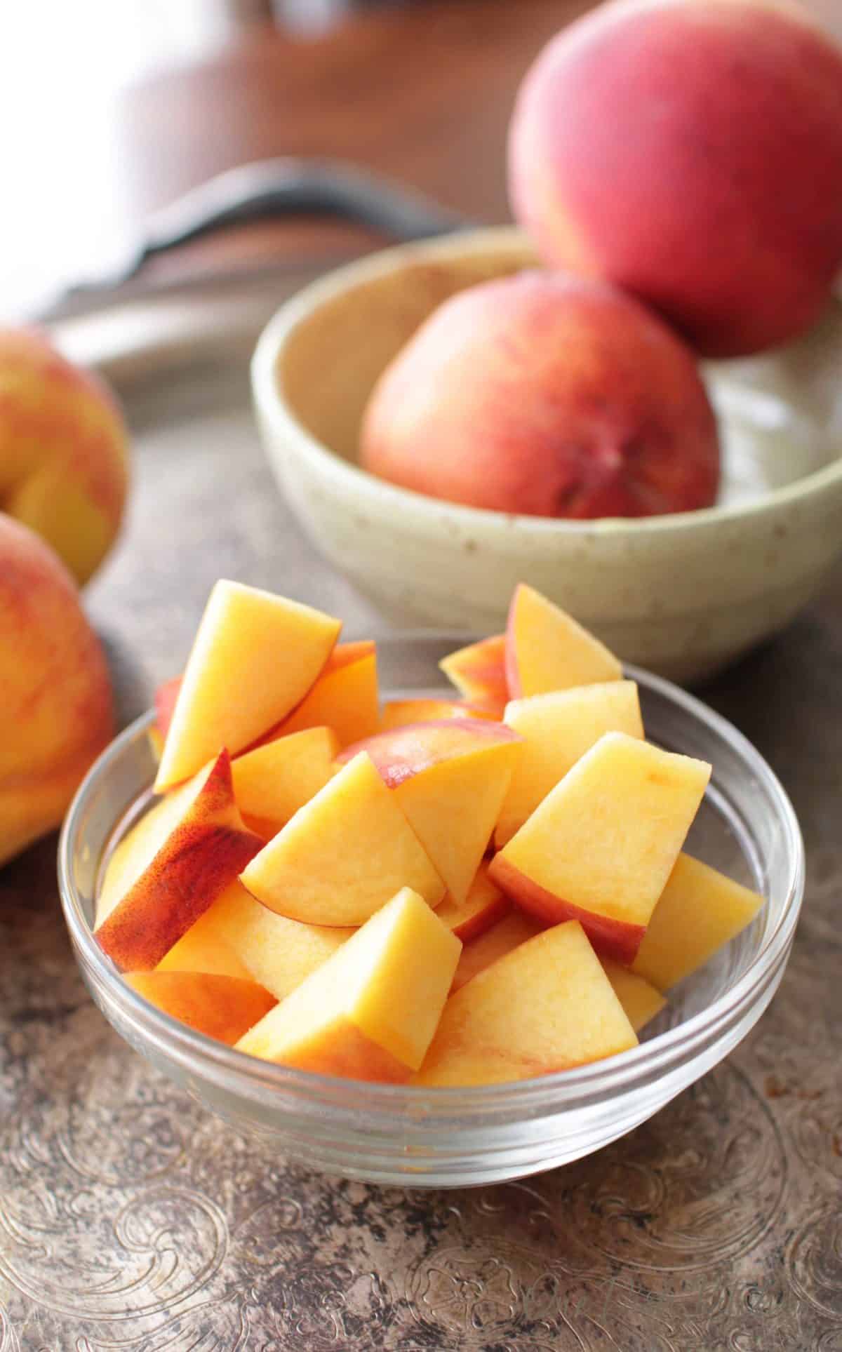 A small bowl filled with chopped peaches next to a bowl of whole peaches on a metal tray.