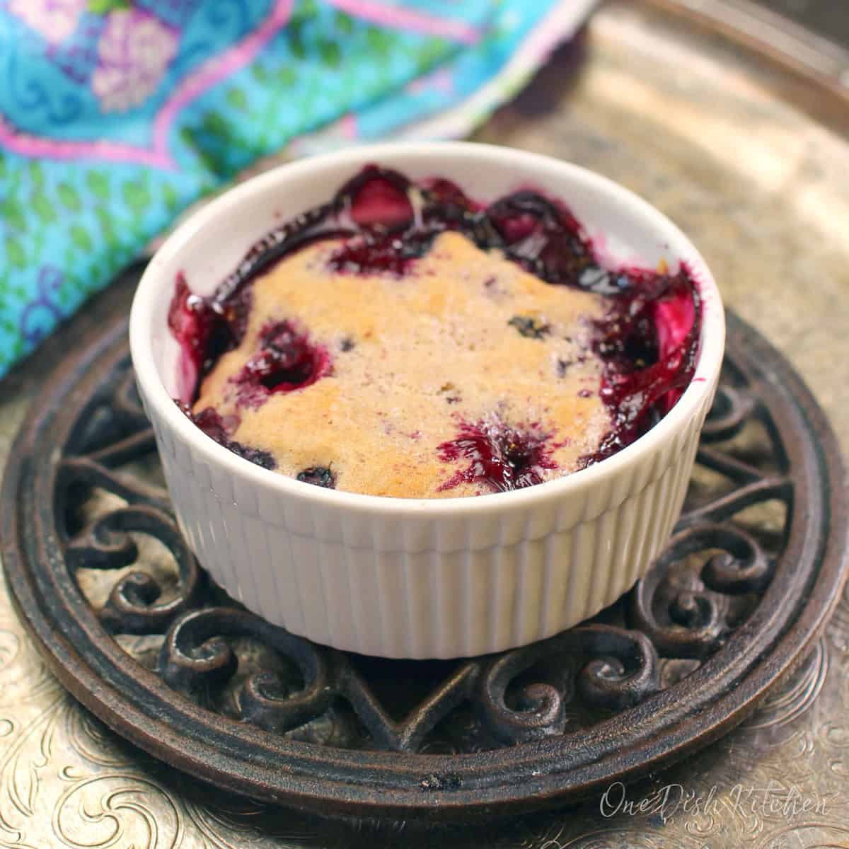 a small blueberry cobbler baked in a white dish on a trivet placed on top of a silver tray.