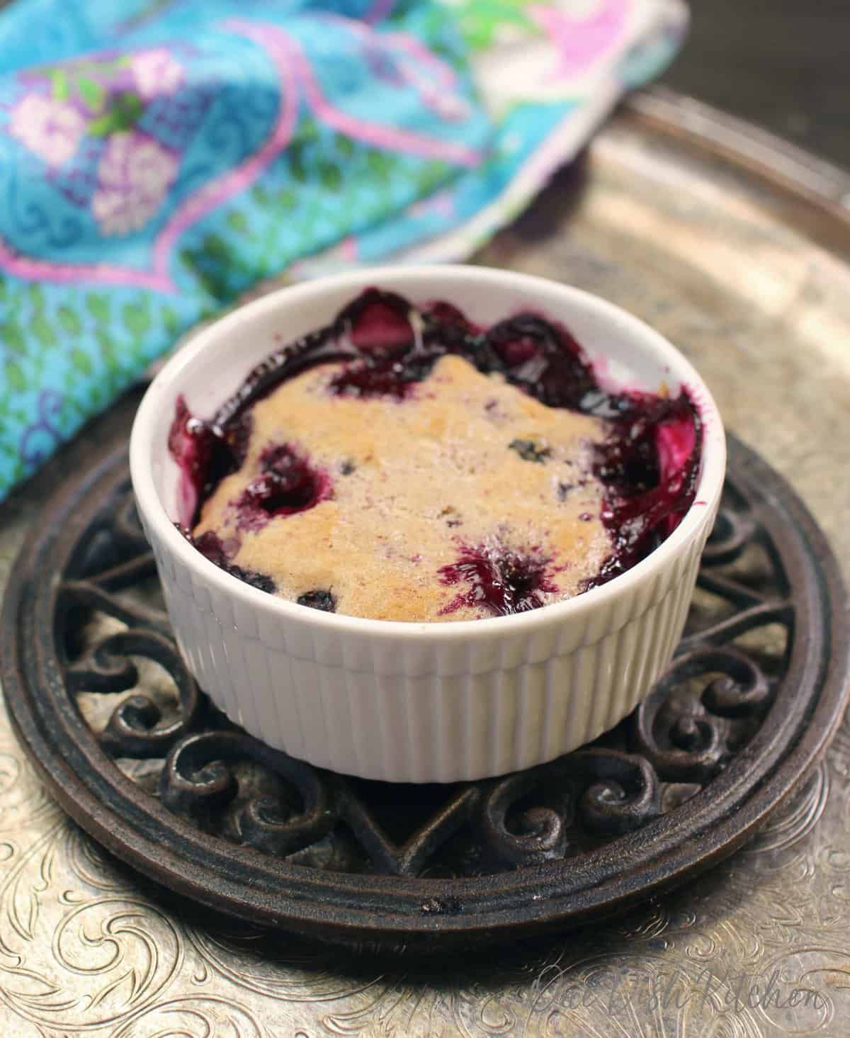 a small blueberry cobbler baked in a white dish on a trivet placed on top of a silver tray.