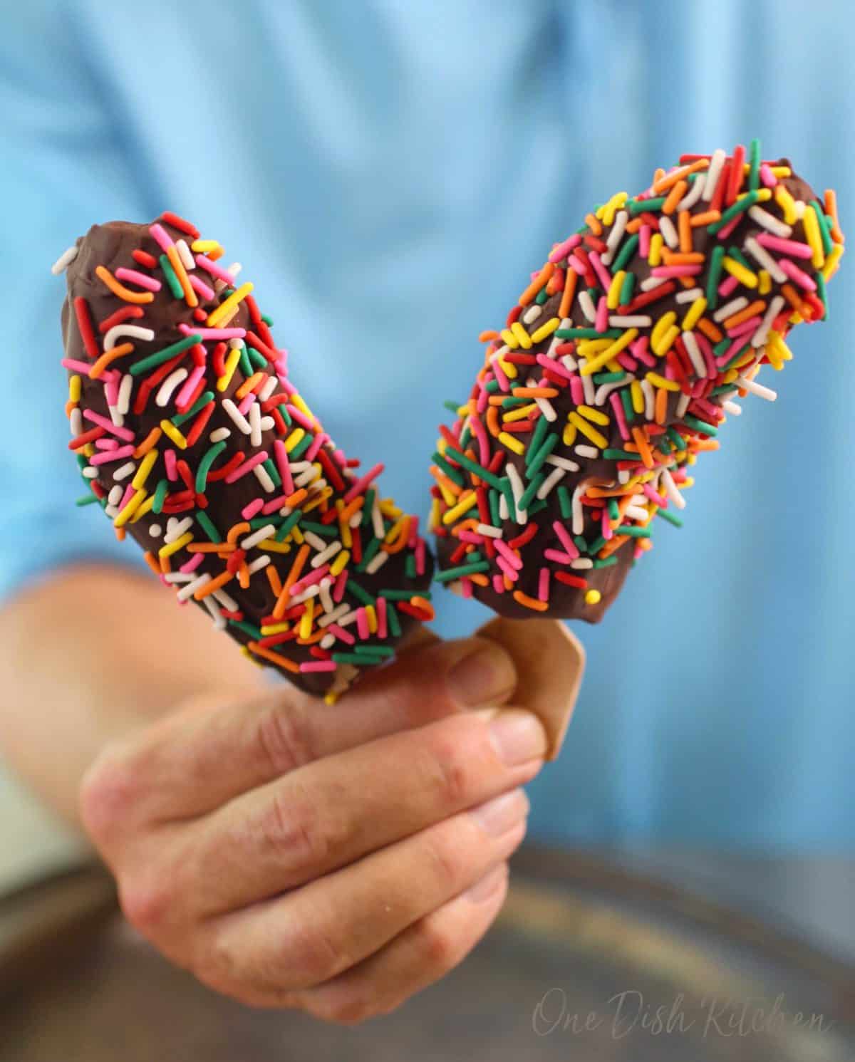 a person holding two frozen chocolate bananas with sprinkles in one hand.