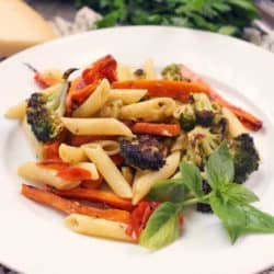 a white plate with penne pasta and roasted broccoli, carrots, and tomatoes on top.