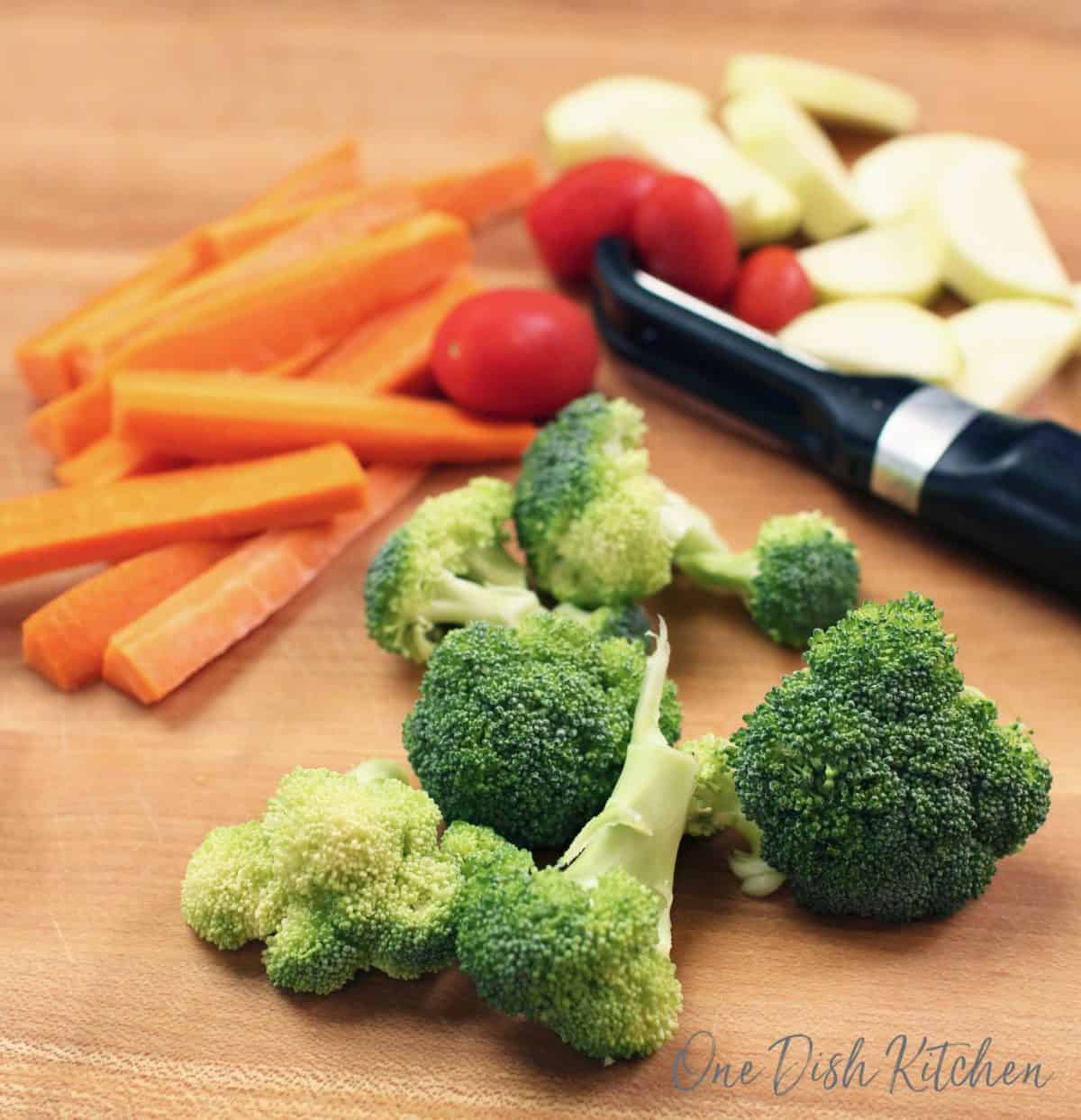 broccoli, carrots, tomatoes and zucchini on a brown cutting board next to a vegetable peeler.