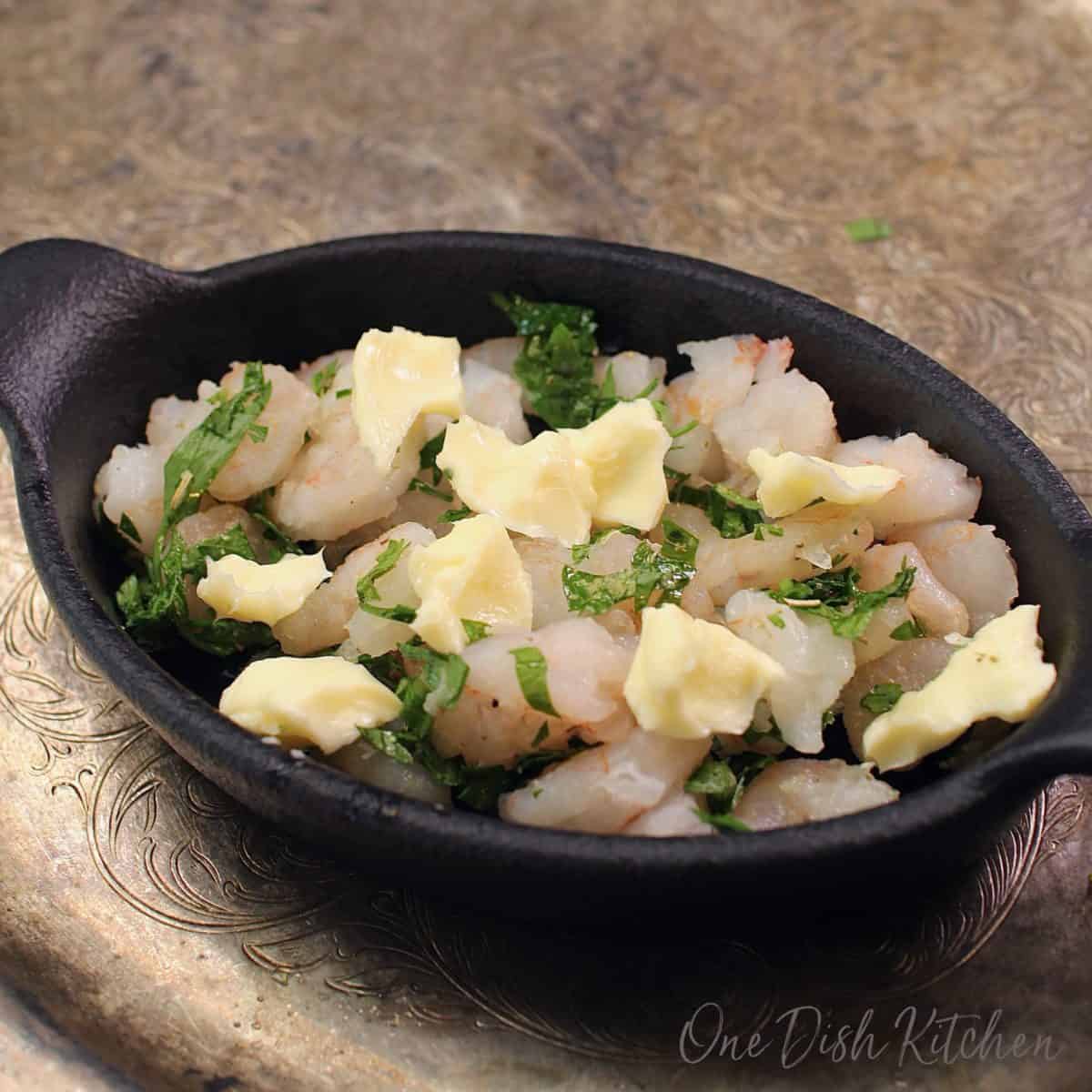 pieces of butter scattered on top of a dish filled with raw shrimp and chopped parsley.