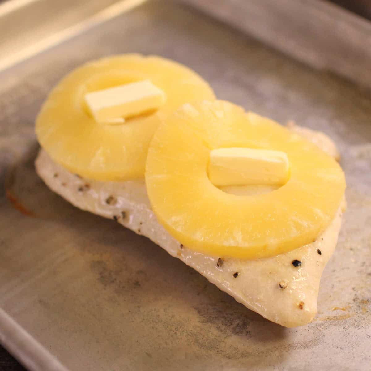 a chicken breast on a baking sheet topped with pineapple slices and butter.