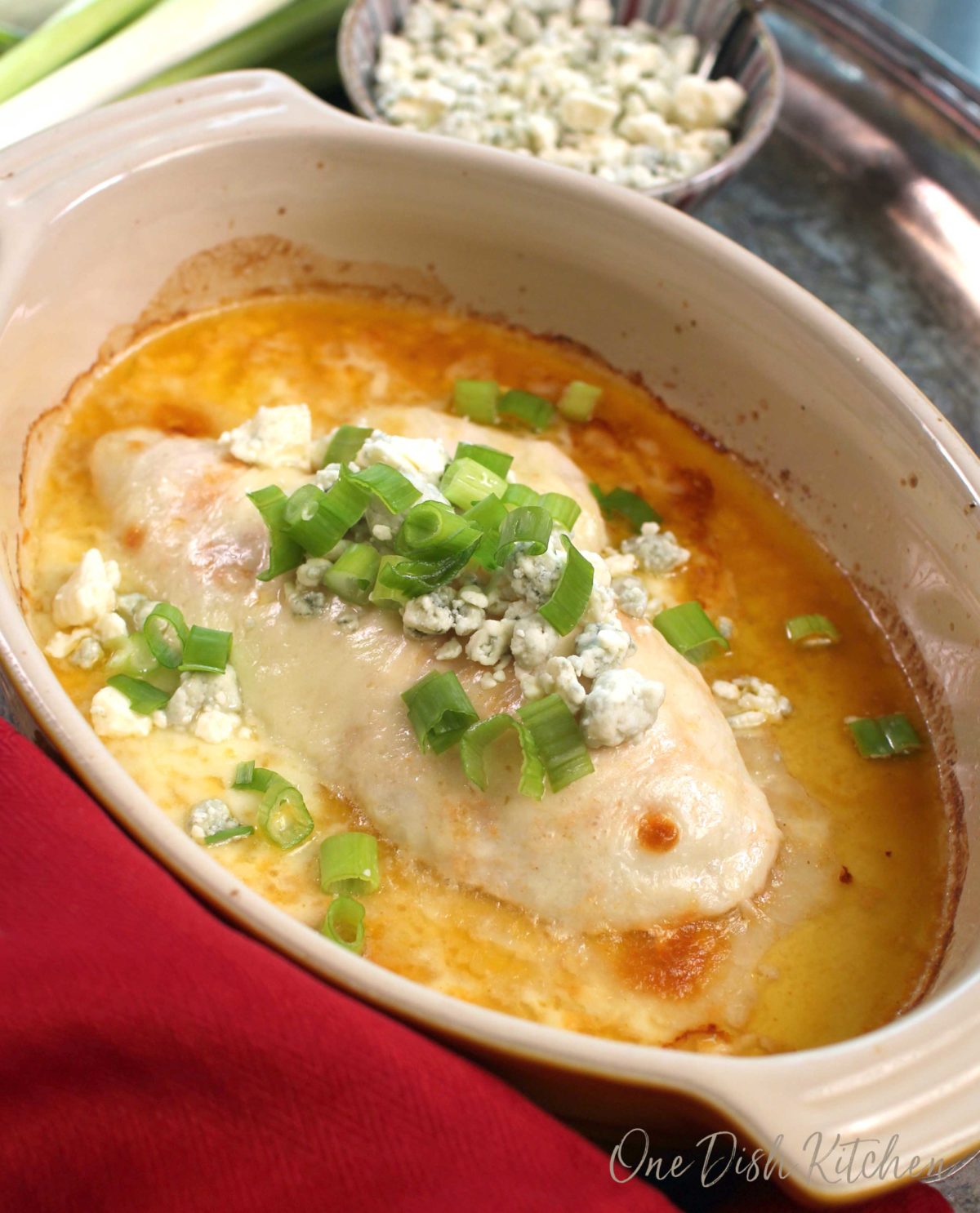 a chicken breast in an oval baking dish topped with blue cheese crumbles and green onions.