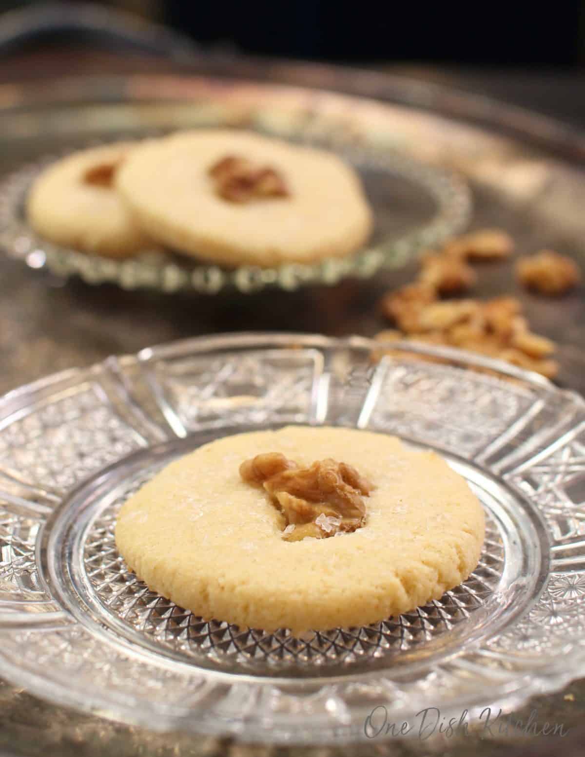 one cookie with a walnut in the center on a clear plate with two cookies in the background.