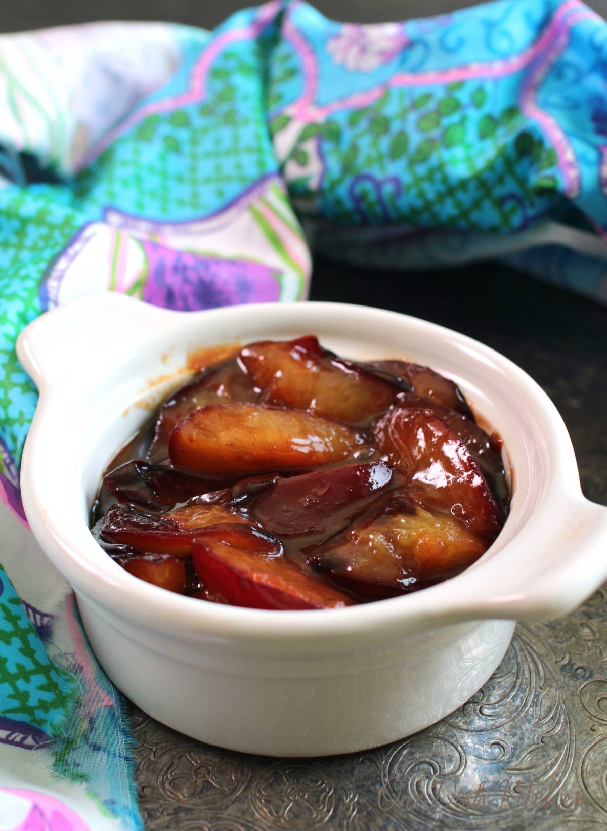 a white bowl filled with softened plums in a sauce next to a aqua colored towel.