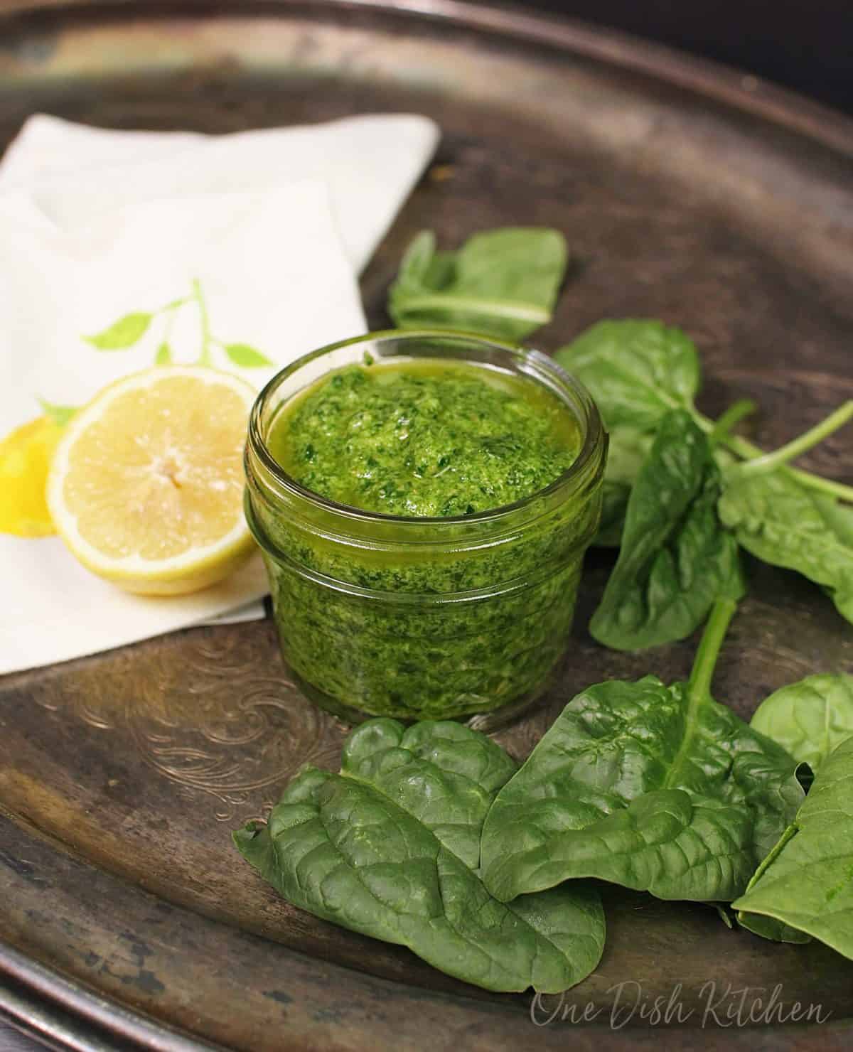 a small jar of pesto on a silver tray next to spinach leaves and a half of a lemon.