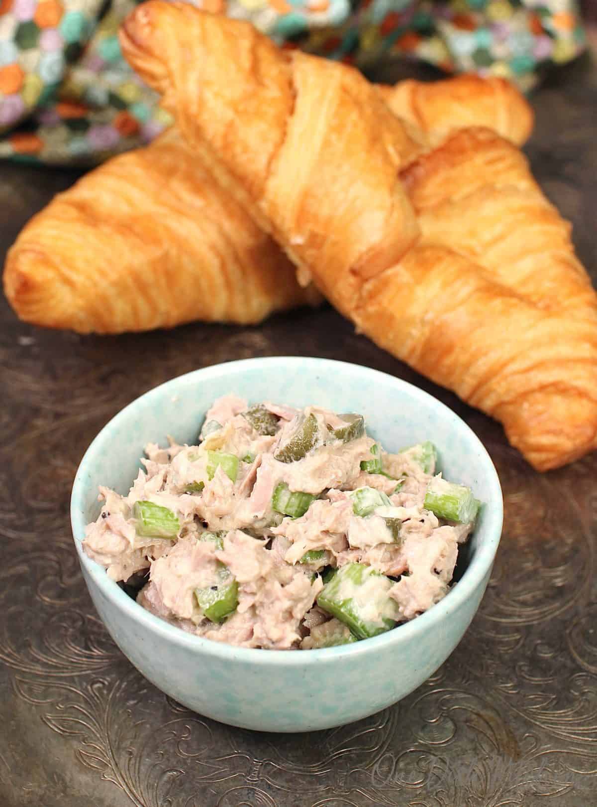 a blue bowl filled with tuna salad next to two croissants on a silver tray.