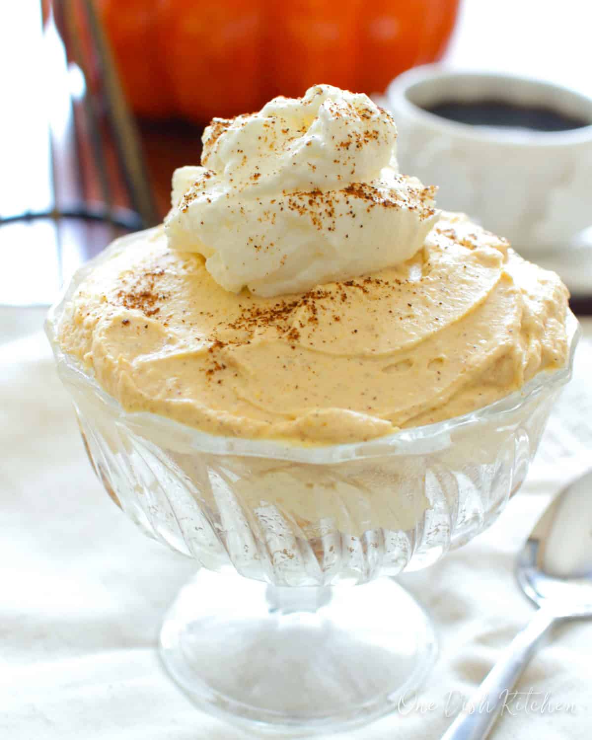 a clear dessert dish with pumpkin cheesecake topped with whipped cream.