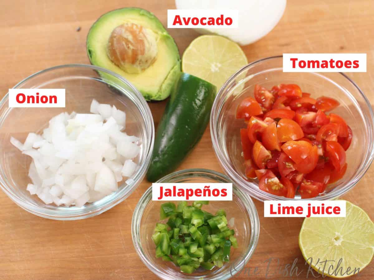 ingredients in pico de gallo on a brown table including onions, avocado, peppers, tomatoes and limes