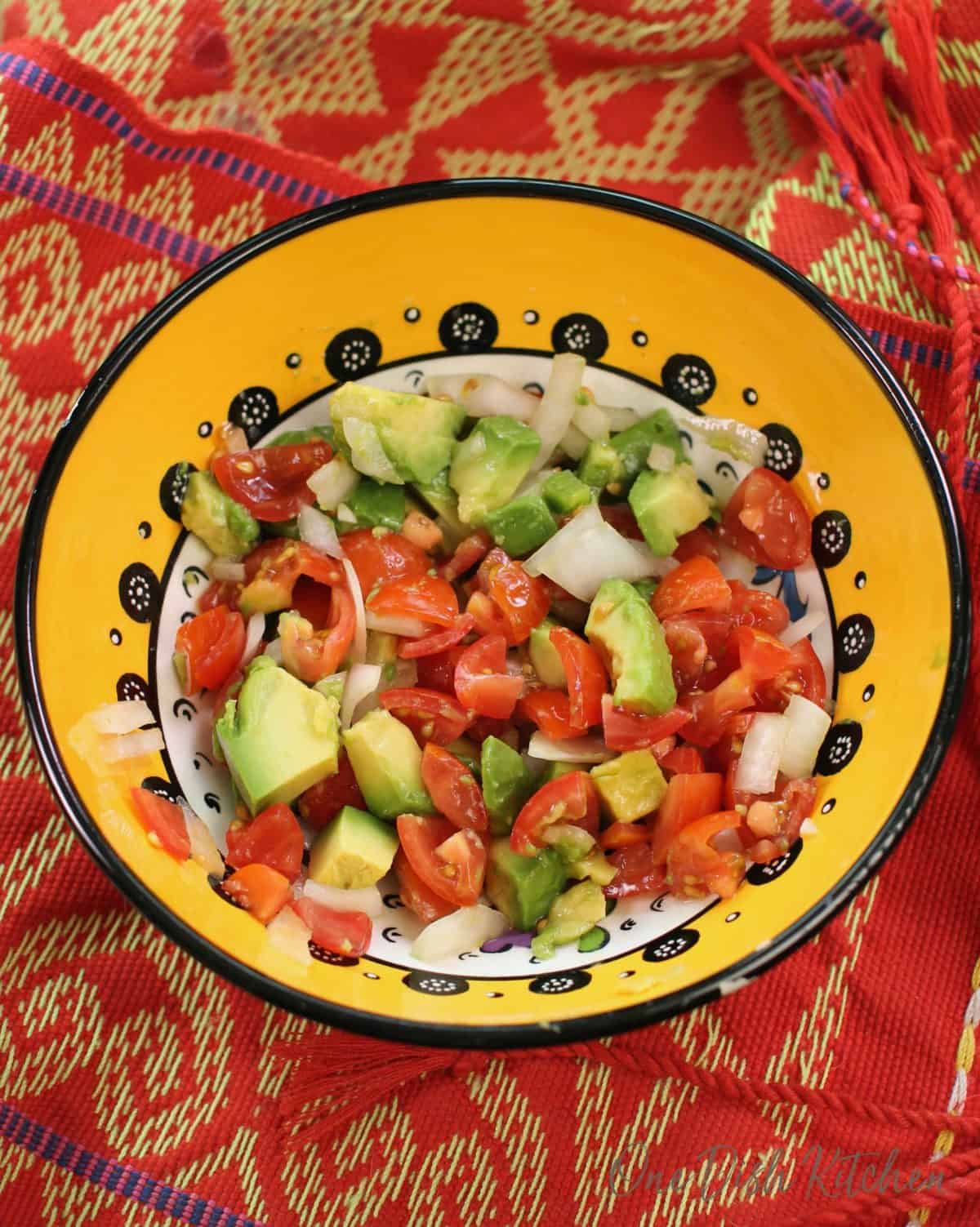 an overhead view of a yellow bowl filled with pico de gallo and chopped avocados on a red and gold towel.