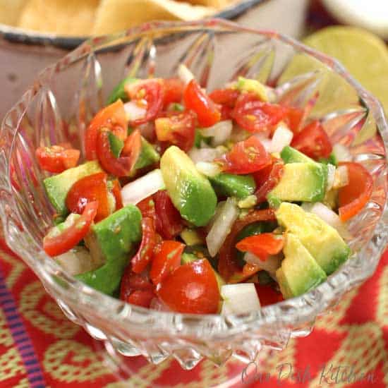 a bowl of pico de gallo with chopped avocados on top surrounded by a red and yellow tablecloth.