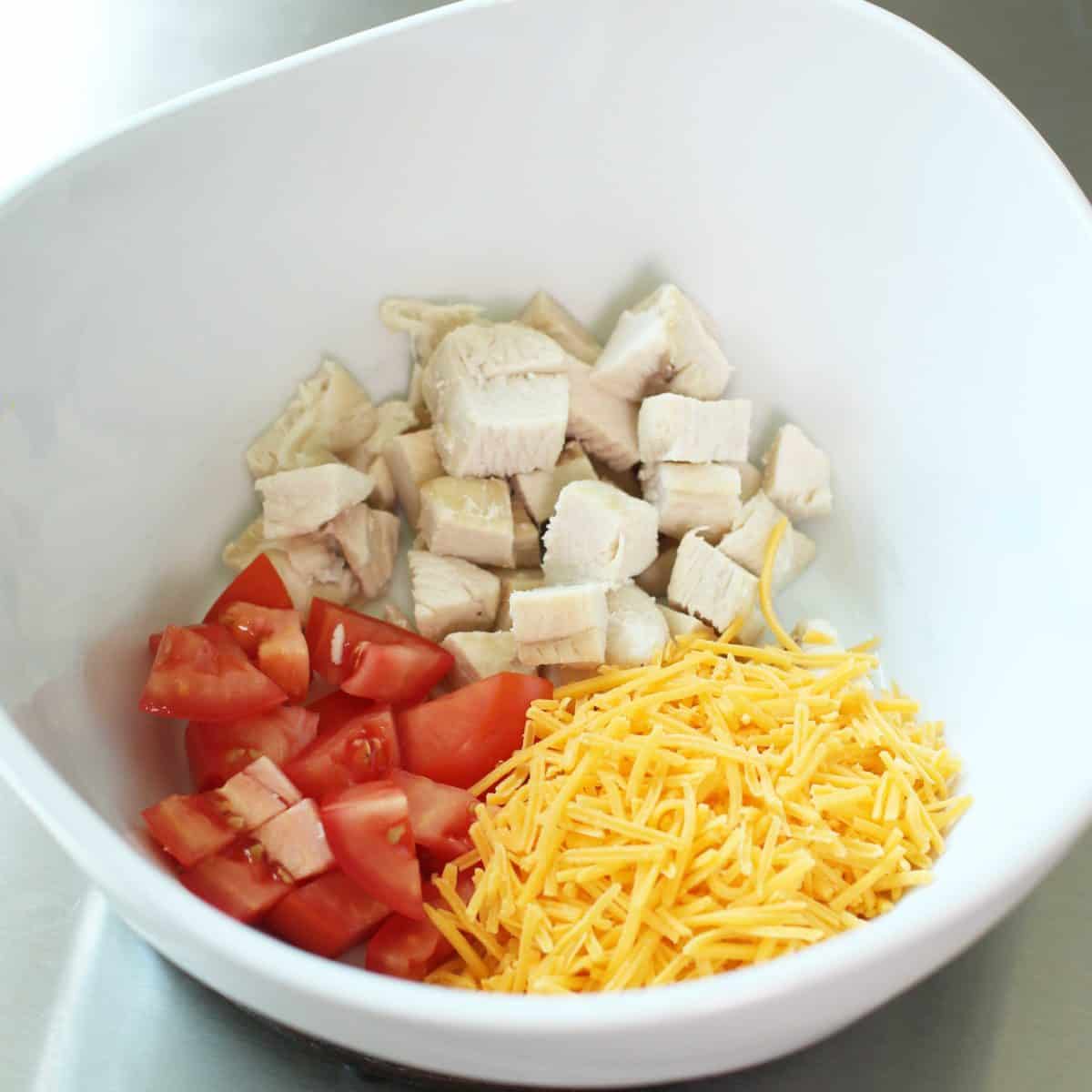 a white bowl filled with chopped chicken, tomatoes, and shredded cheddar cheese.