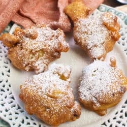 four crispy apple fritters topped with powdered sugar on a white plate.