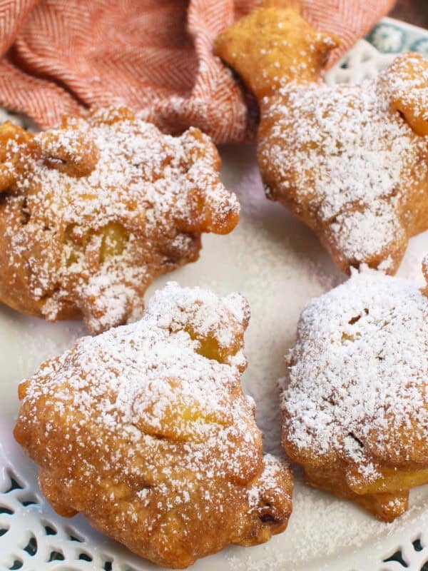 four crispy apple fritters topped with powdered sugar on a white plate.