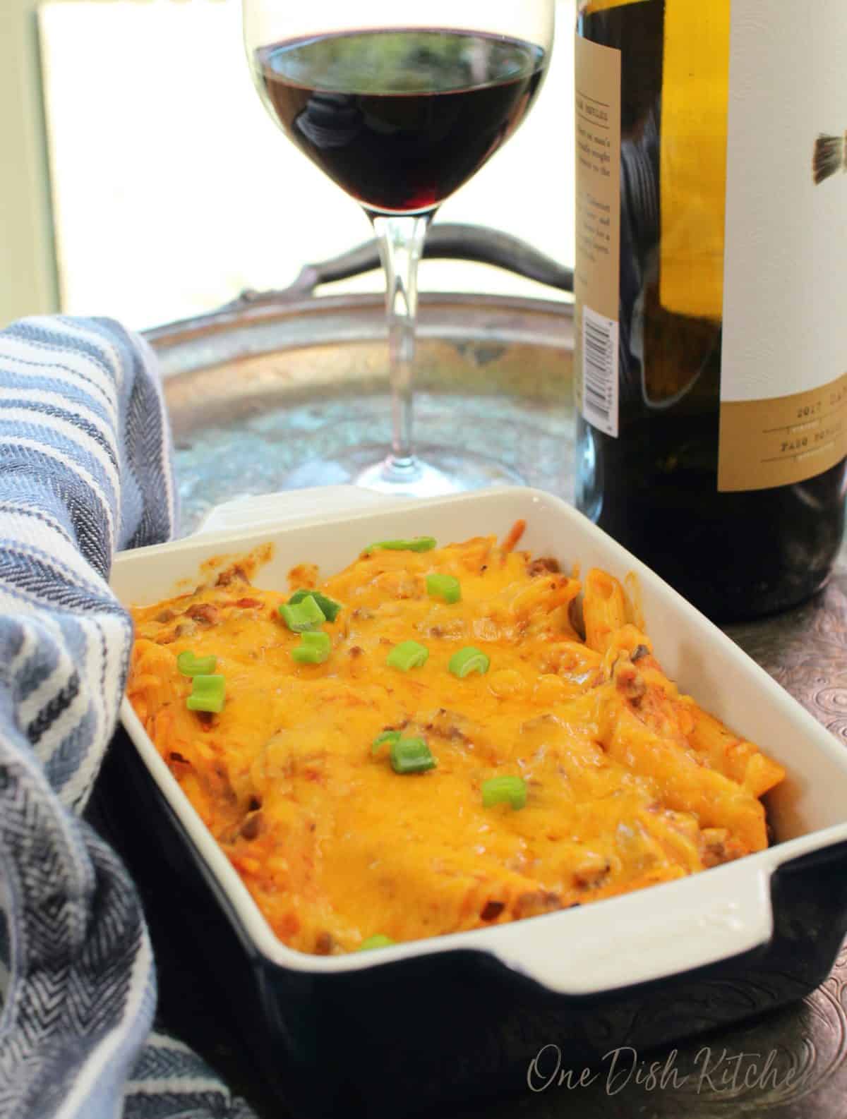 A single serving hamburger casserole in a blue baking dish next to a glass of red wine.