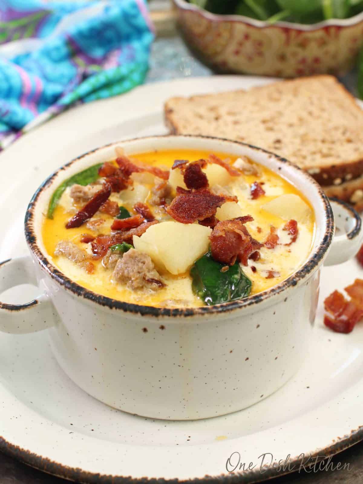 a small bowl of soup filled with potatoes, bacon, sausage and spinach on a plate next to two slices of bread.