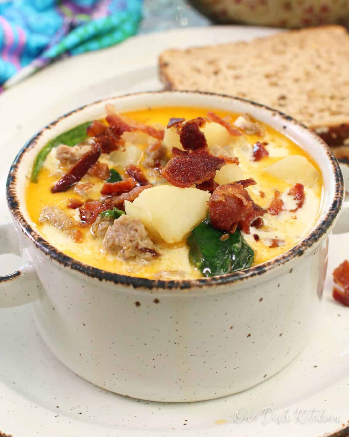 a small bowl of soup filled with potatoes, bacon, sausage and spinach on a plate next to two slices of bread.