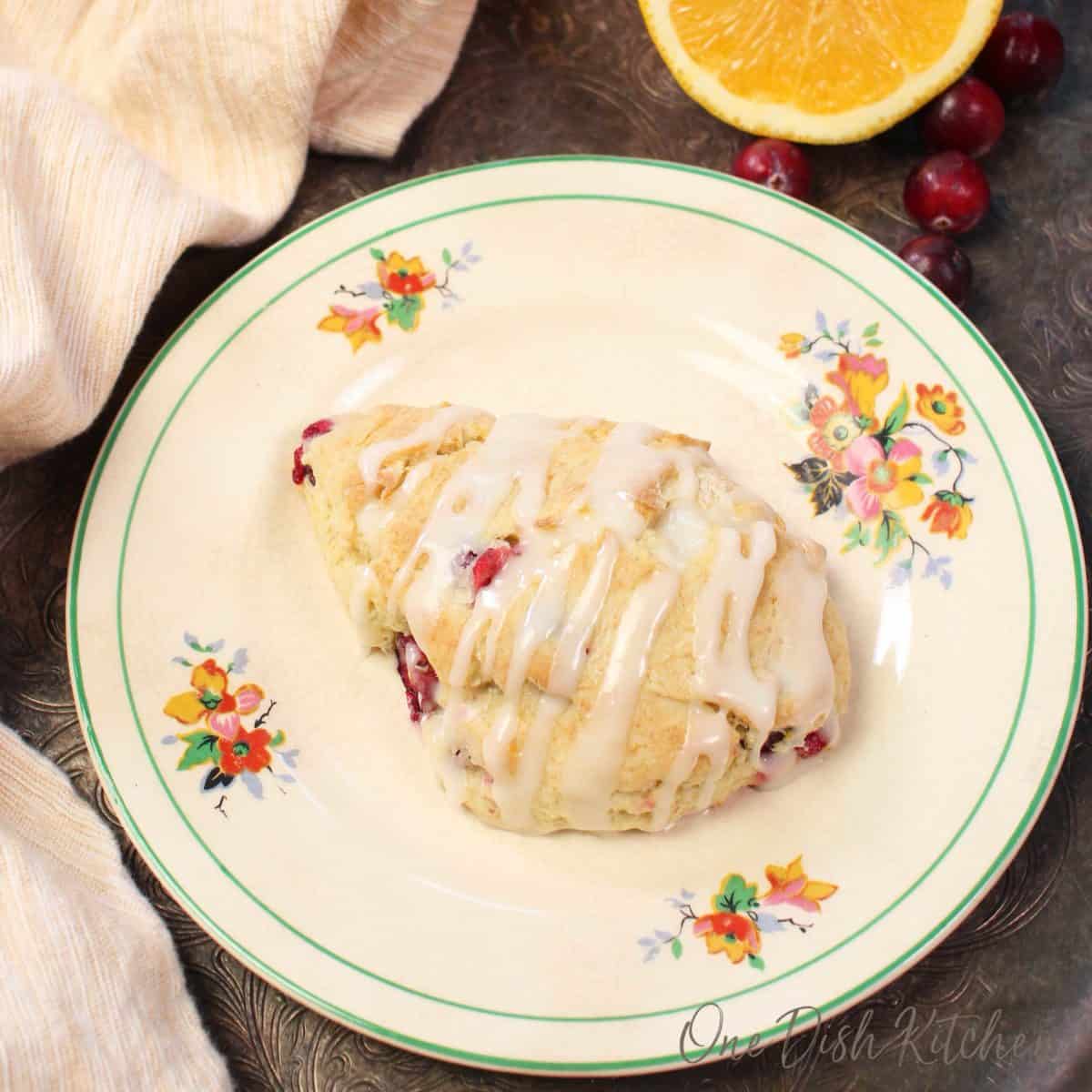 one scone on a flowered plate next to fresh cranberries scattered around a silver tray.