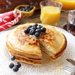 three pancakes on a white plate with blueberries on top and scattered around the plate.