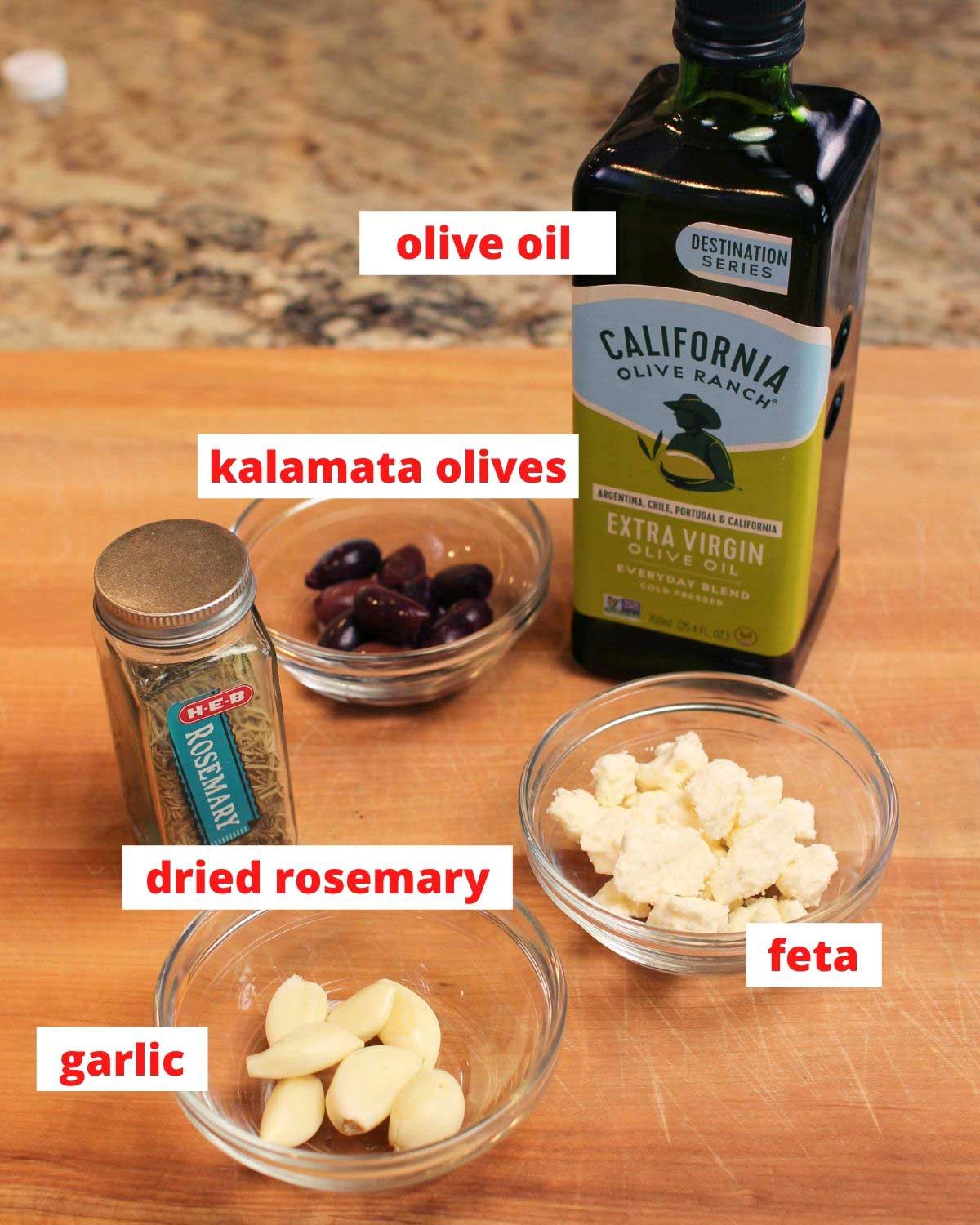 olive oil, olives, spices, cheese and a bowl of garlic cloves on a cutting board.